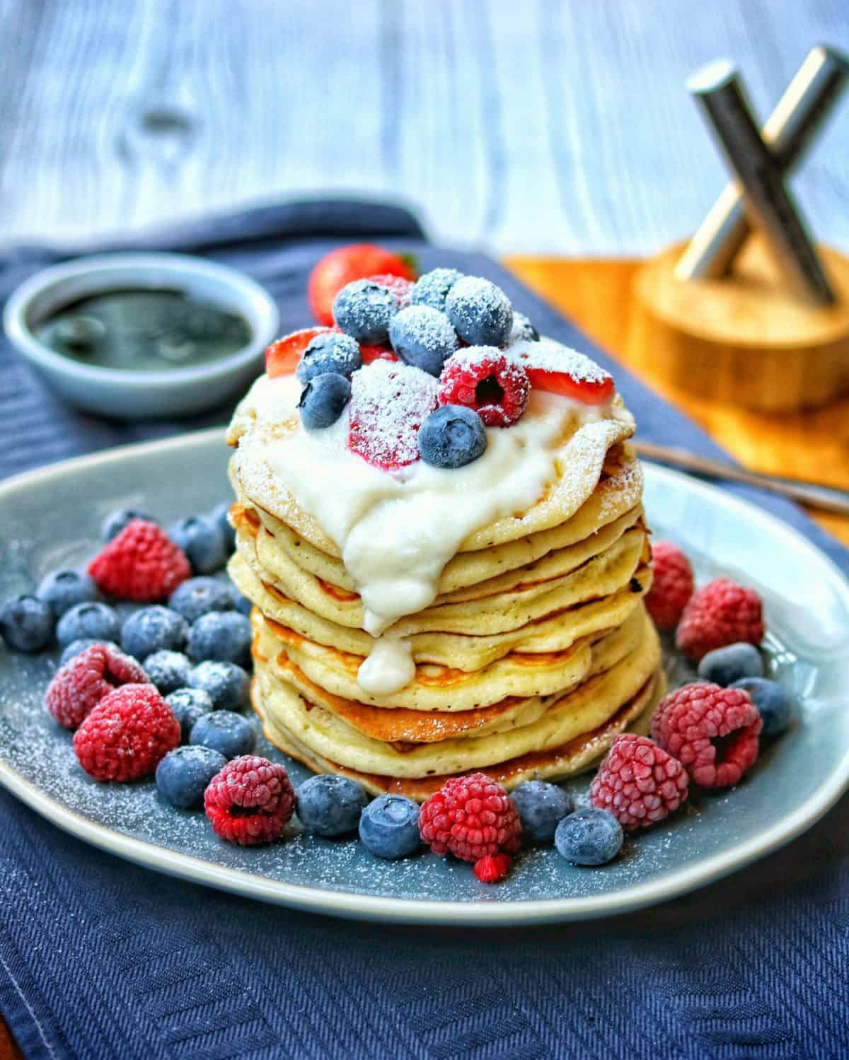 Ein blauer ovaler Teller mit einem Stapel Pancakes. Obendrauf Sahne und Beeren. Außenrum Beeren als Deko.