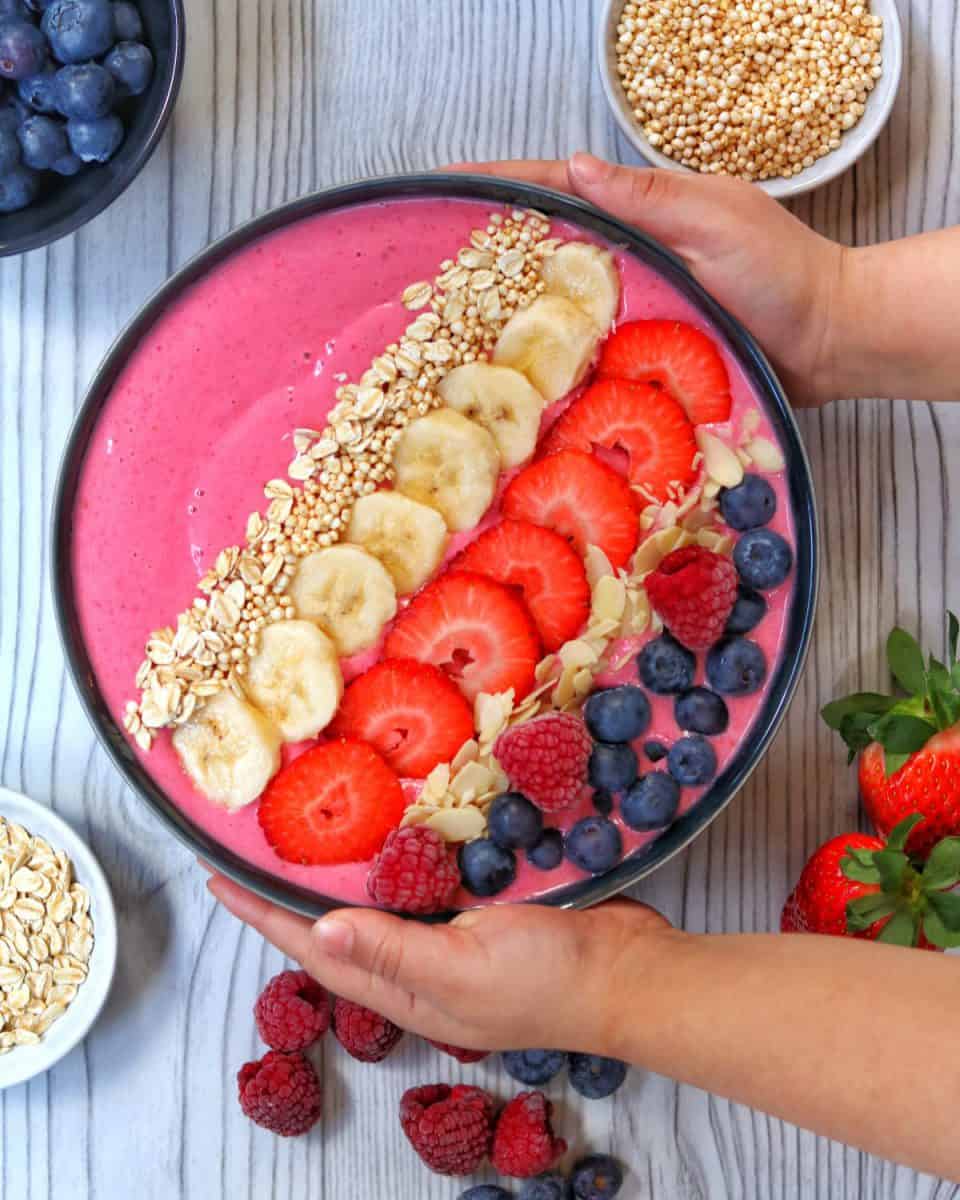 Zwei Hände halten die Smoothie-Bowl. Aussenrum Früchte und Quinoa als Deko.