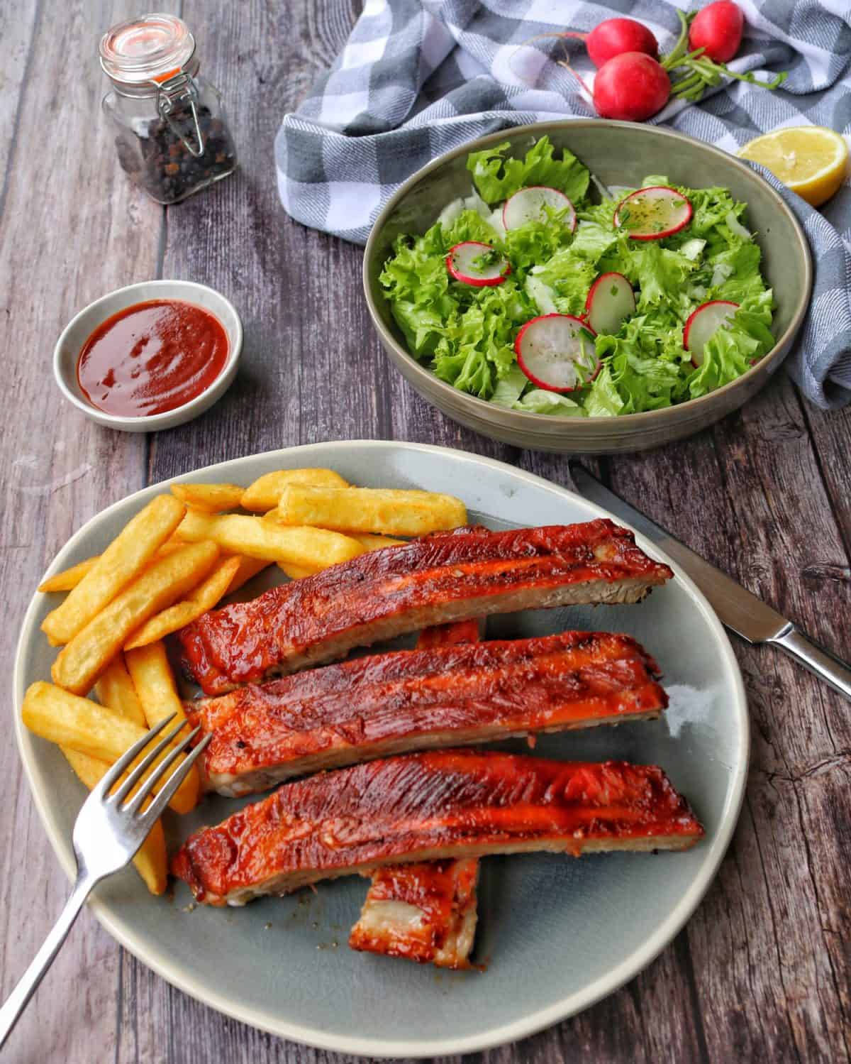 Ein ovaler Teller mit Spareribs und Pommes. Dahinter eine Schale mit Salat und Deko.