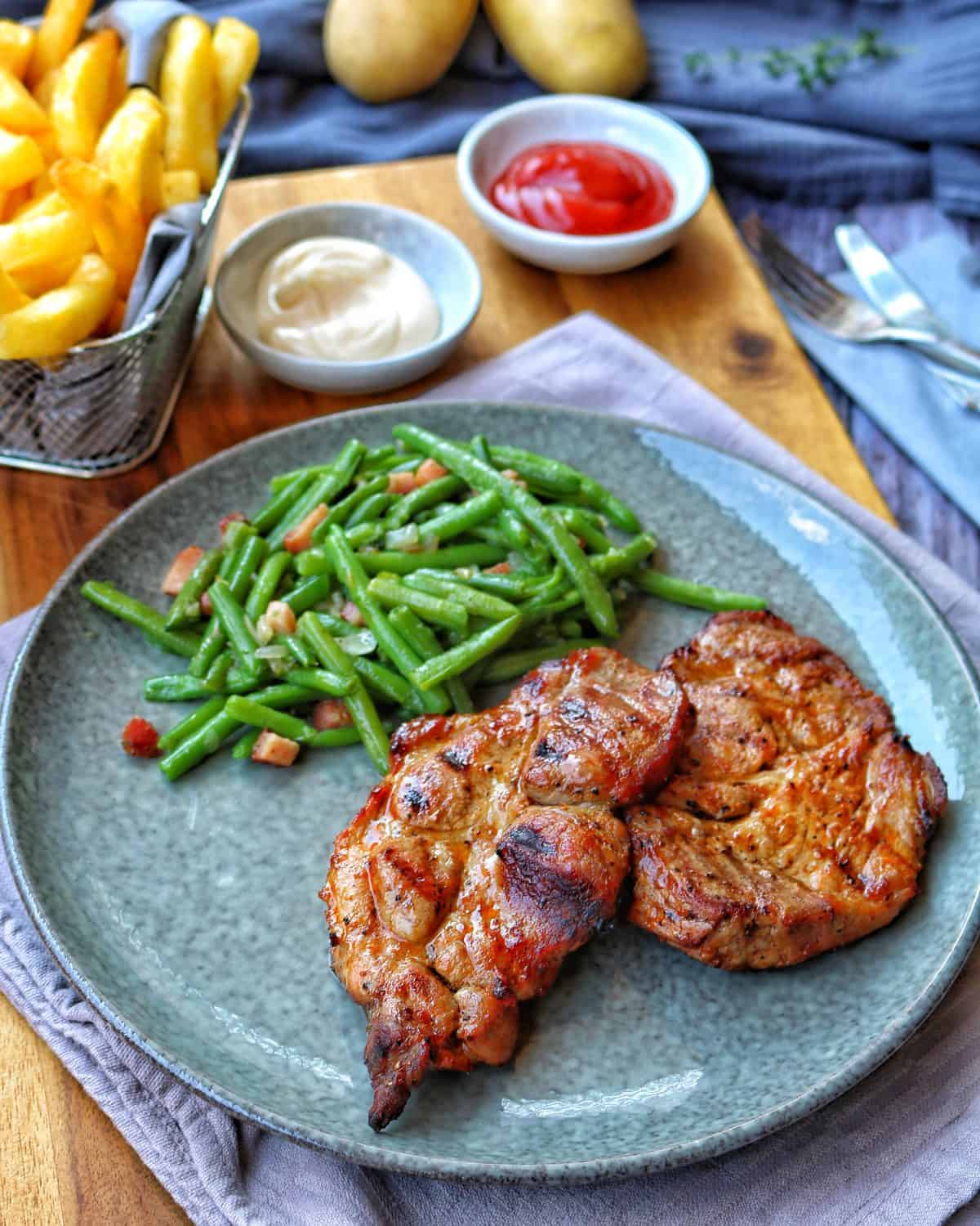 Ein Teller mit Nackensteak mit Speckbohnen und Pommes im Hintergrund