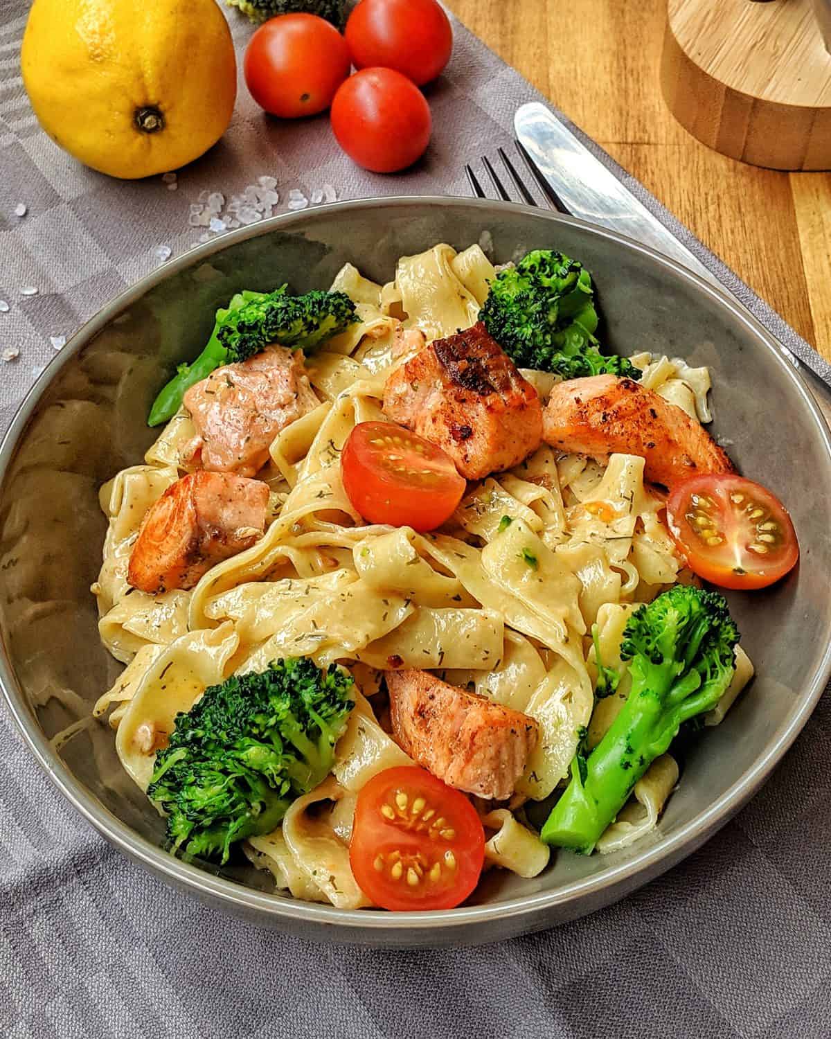 Ein Teller mit Nudeln und Lachs in Sahnesoße. Dabei sind Brokkoli und Tomaten.