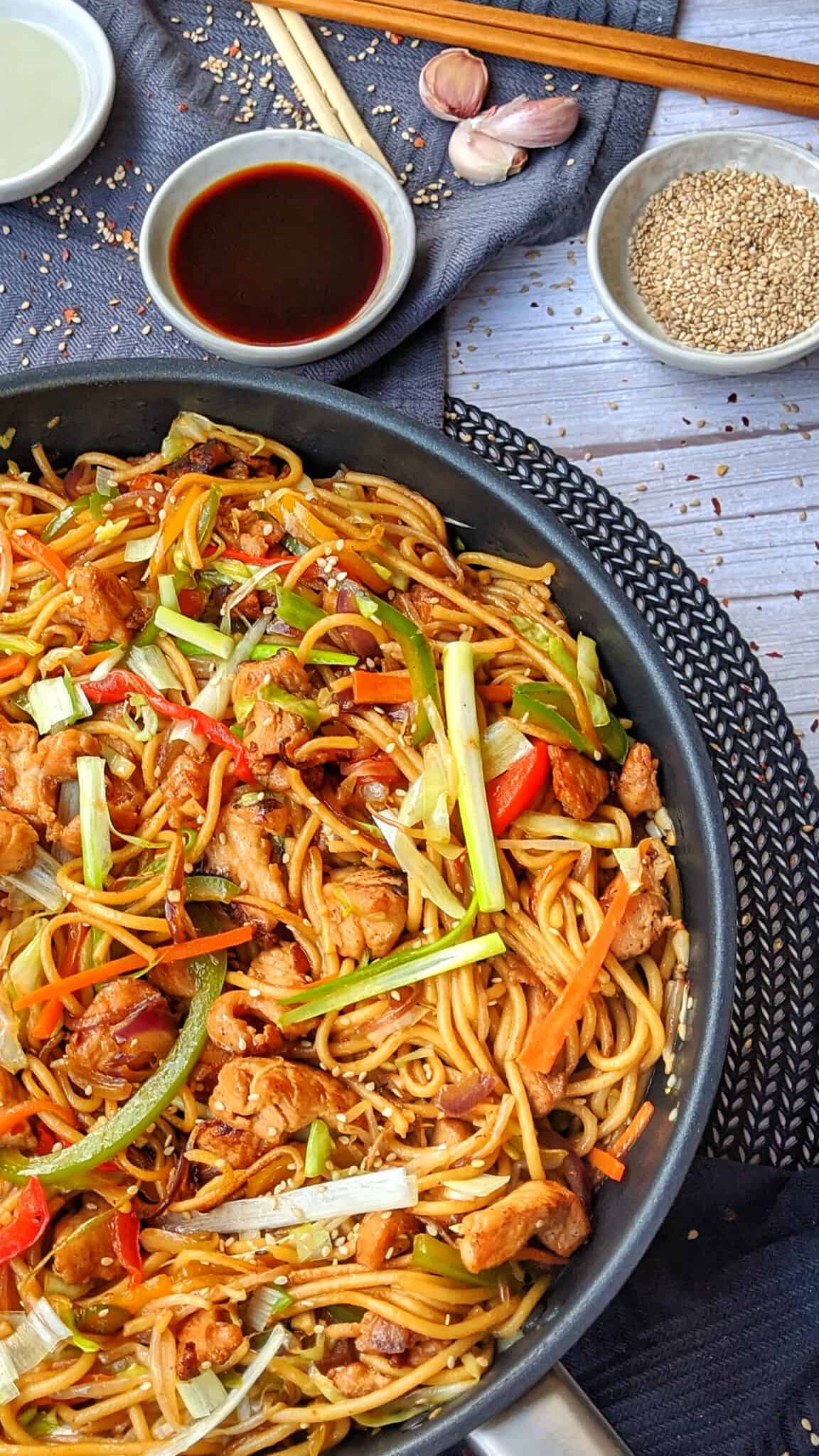A pan with a chicken Chow Mein. Behind it, a small dish with soy sauce and sesame seeds, as well as decoration.