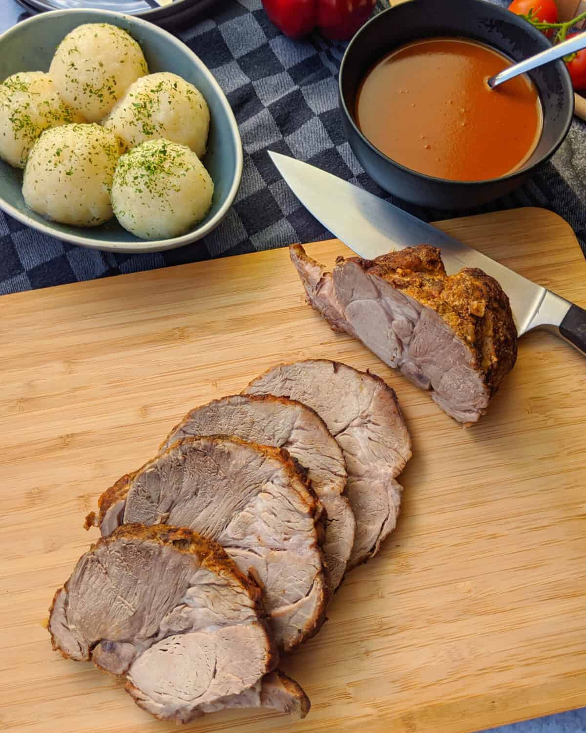 Ein angeschnittener Schweinebraten auf einem Holzbrett. Im Hintergrund Klöße und Bratensoße.