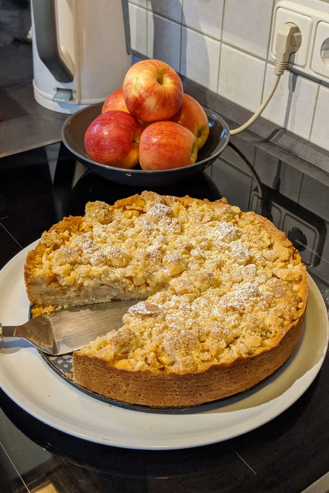 Ein Apfelkuchen mit Streusel auf einer Servierplatte. Im Hintergrund eine Schale mit Äpfeln.