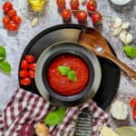 Eine Schüssel mit Tomatensauce. Aussen herum Tomaten, Basilikum, Parmesan Salz, Olivenöl und Knoblauch als Deko.