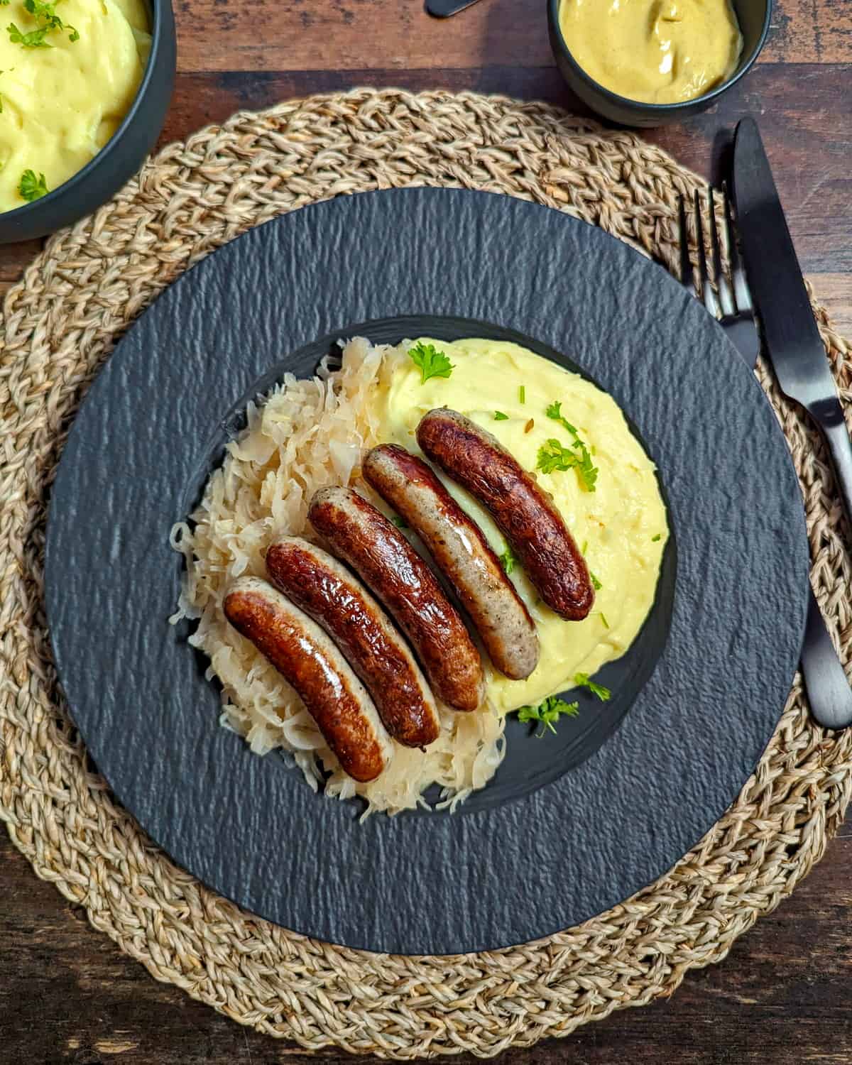 Ein schwarzer Teller mit Nürnberger Rostbratwürstchen mit Sauerkraut und Kartoffelpüree.