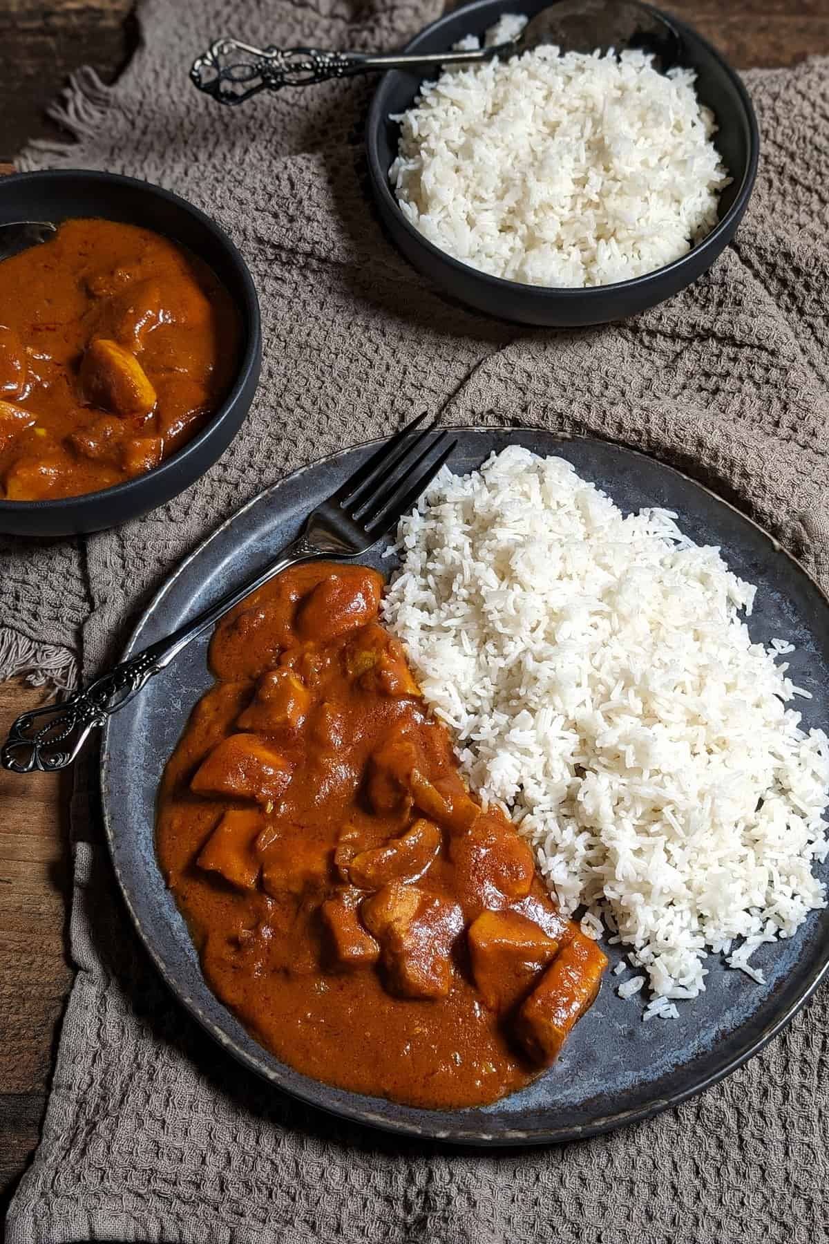 Ein dunkler Teller mit Hähnchen-Curry und Reis. Darüber eine Schale mit Hähnchen-Curry und eine Schale mit Reis.