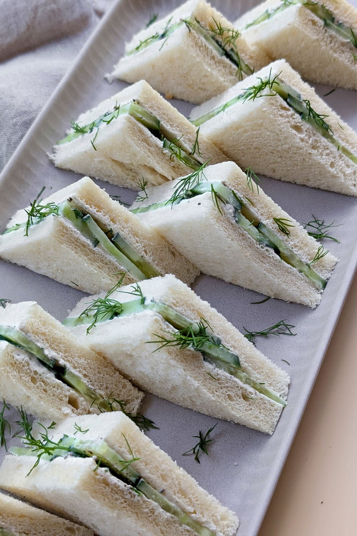 Eine Nahaufnahme einer rechteckigen Servierplatte mit dreieckförmigen Gurken-Sandwiches.