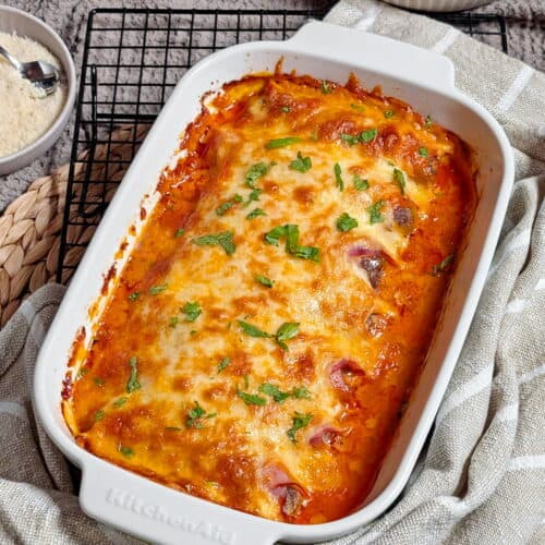 Eine weiße Auflaufform mit Käse überbackenen Schinken-Hack-Röllchen in Tomatensoße.