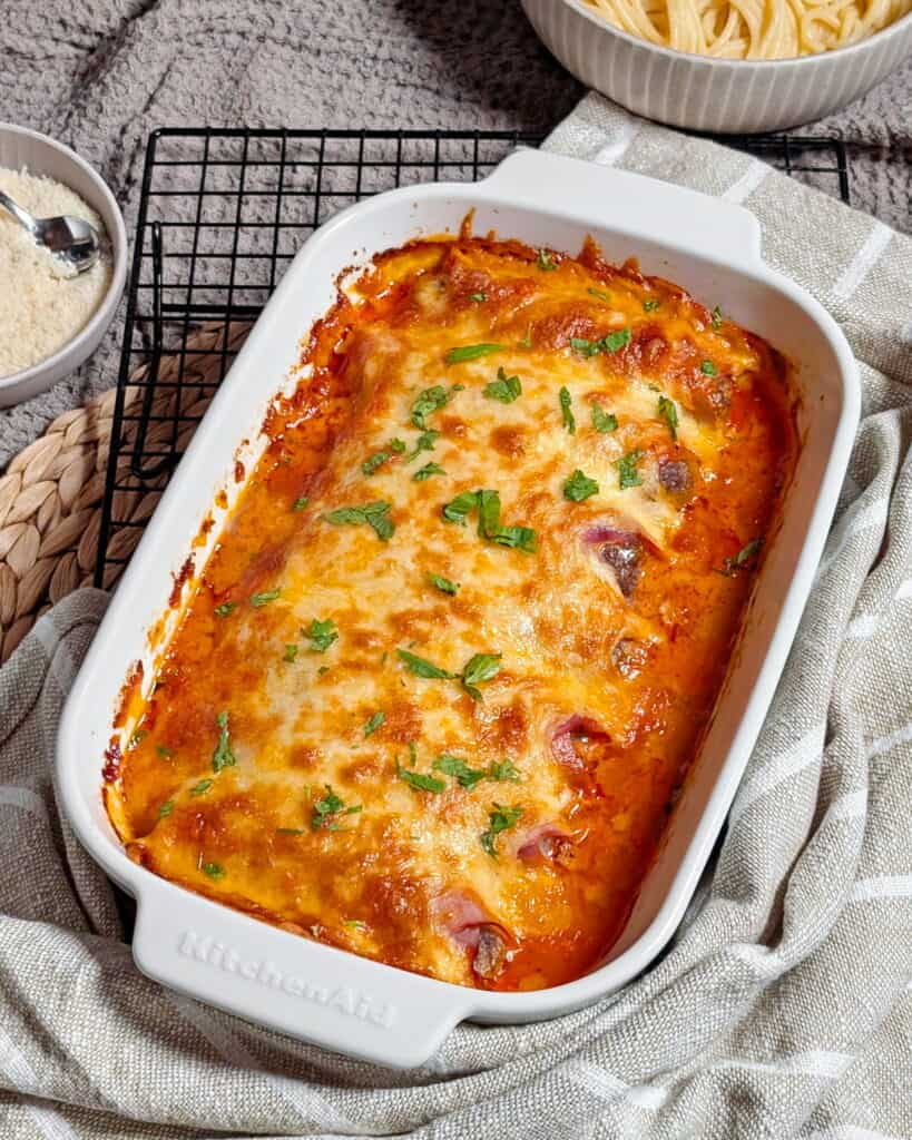 Eine weiße Auflaufform mit Käse überbackenen Schinken-Hack-Röllchen in Tomatensoße.